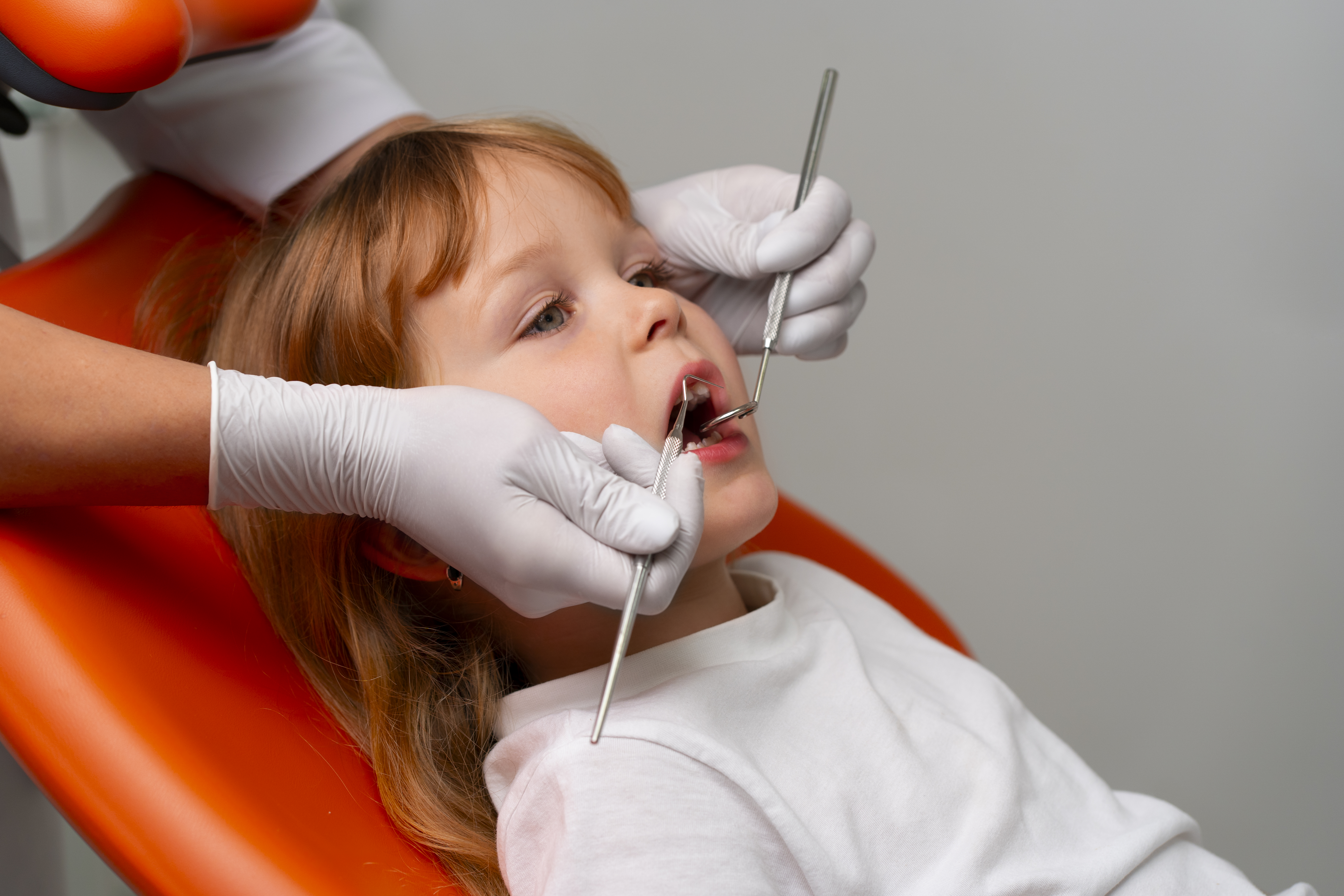 Pediatric dentist examining young child’s teeth with dental mirror and probe during routine checkup in modern clinic chair