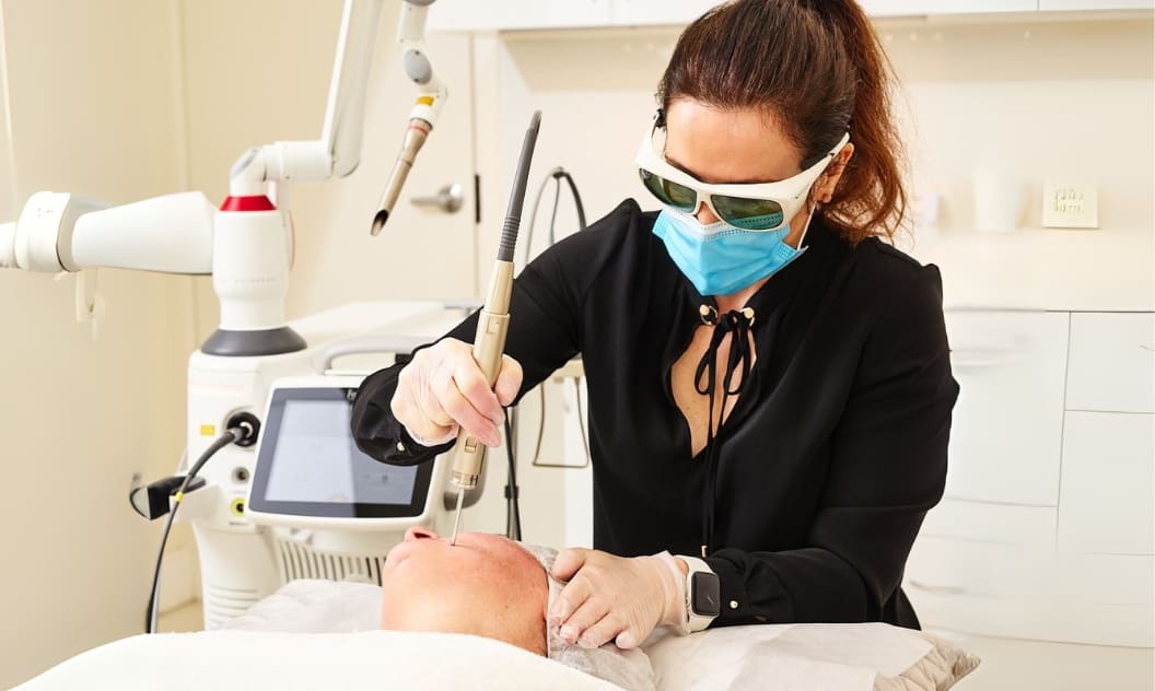 The Benefits of Our Aesthetic Laser Systems A masked specialist performs a laser facial treatment on a patient using advanced aesthetic equipment.