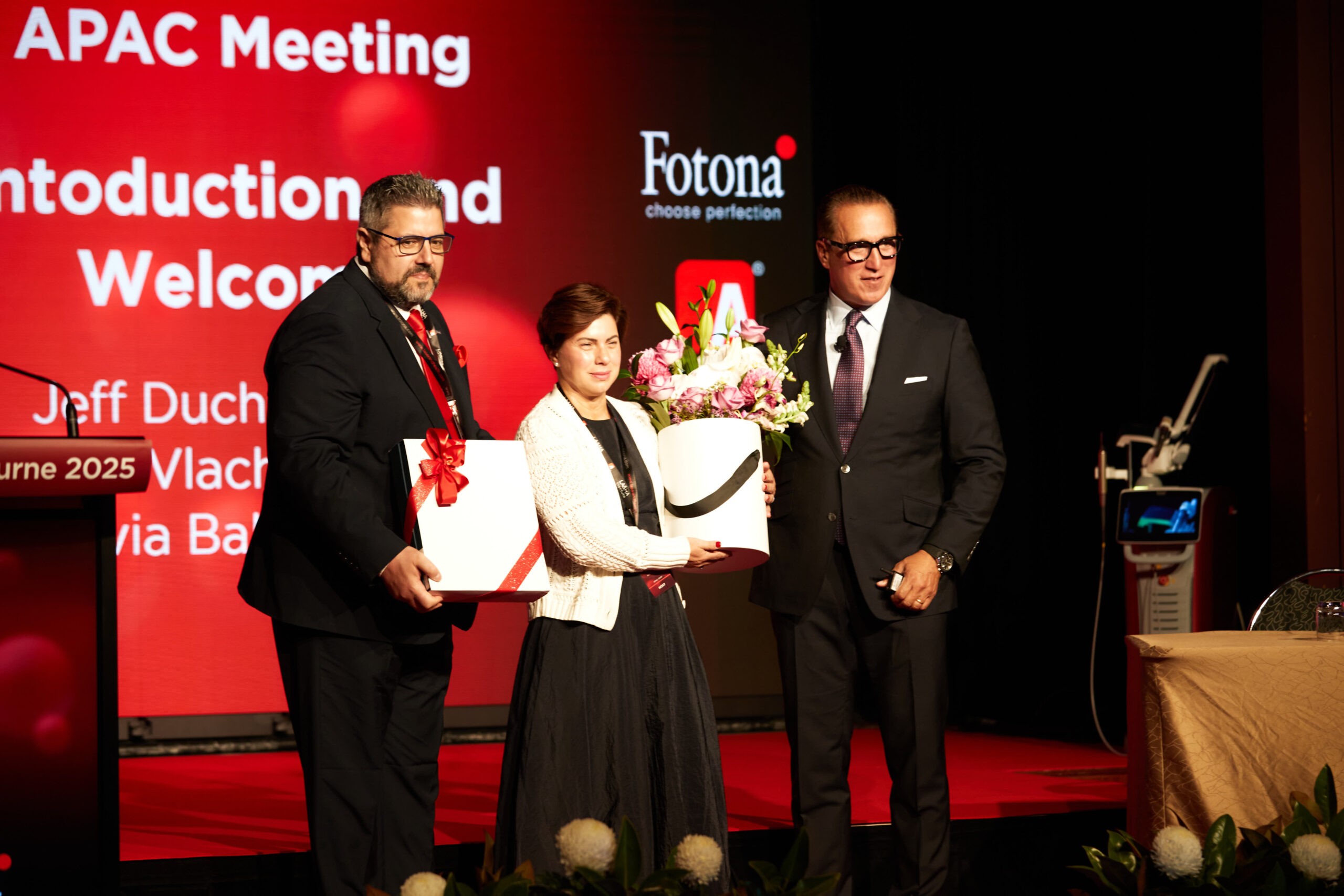 Sam, Sylvia, Jeff-min Award presentation at Fotona APAC Meeting with speakers on stage holding gift box and bouquet during conference ceremony